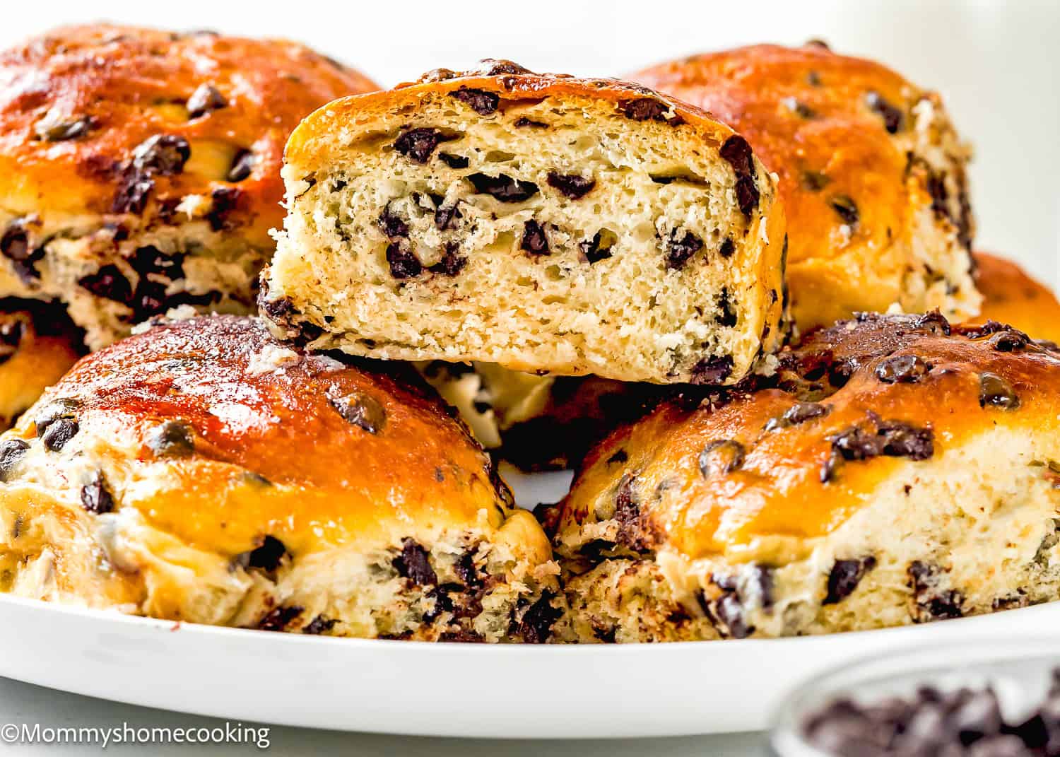 A plate of golden brown Eggless Chocolate Chip Brioche Rolls, with one roll cut in half to reveal the soft, chocolate-filled interior.