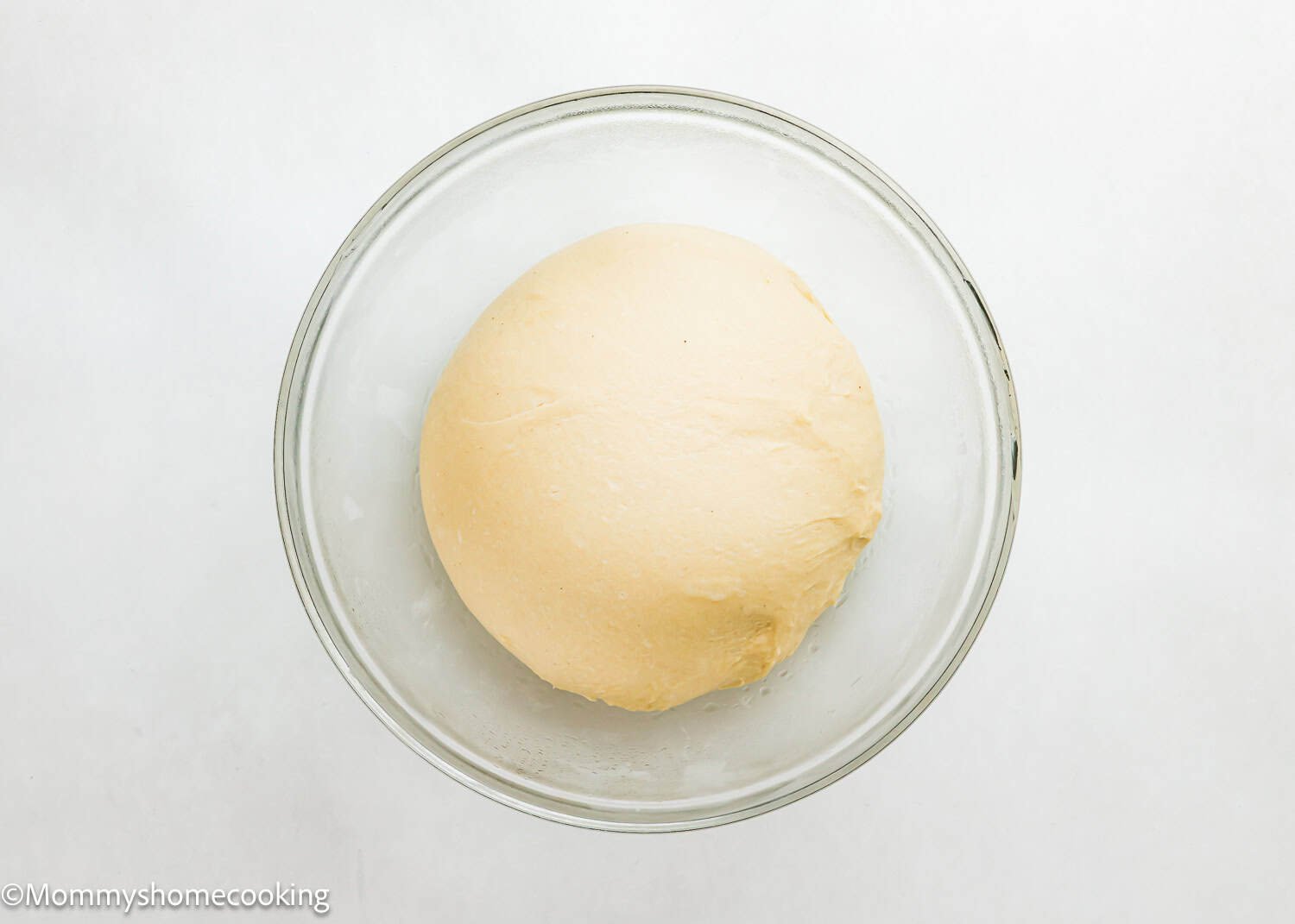 A ball of dough for Eggless Chocolate Chip Brioche Rolls sits in a clear glass bowl on a white surface.