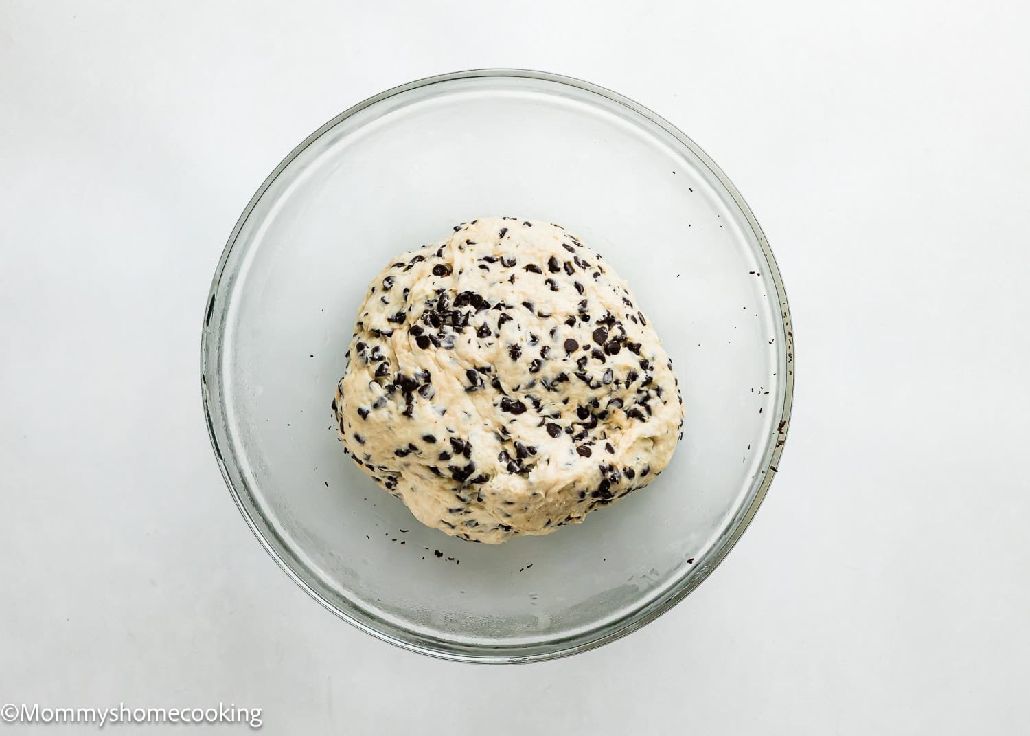 A glass bowl containing a ball of dough for eggless chocolate chip brioche rolls sits on a white surface.