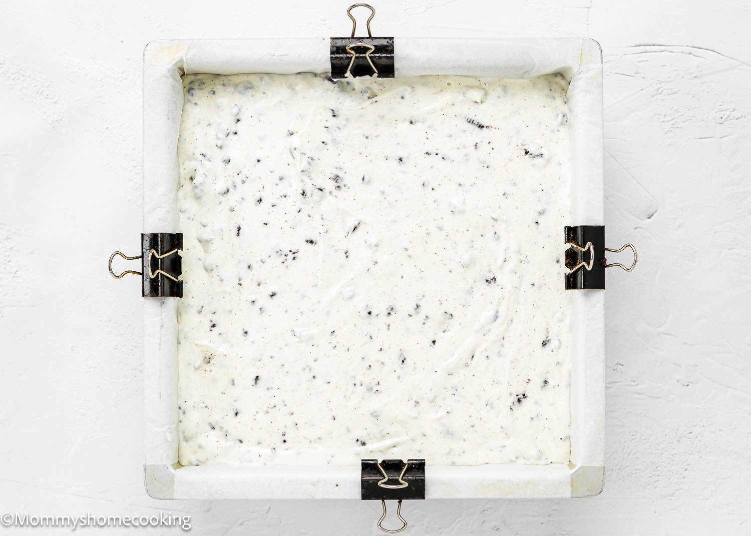 A square baking pan lined with parchment paper and secured with binder clips, filled with a creamy, speckled eggless ice cream cake mixture ready to be baked.