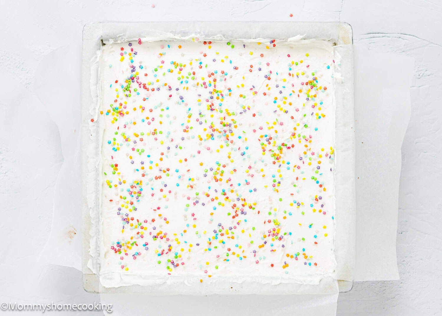 An Eggless Ice Cream Cake, square-shaped with white frosting and topped with colorful round sprinkles, sits in a parchment-lined baking pan.