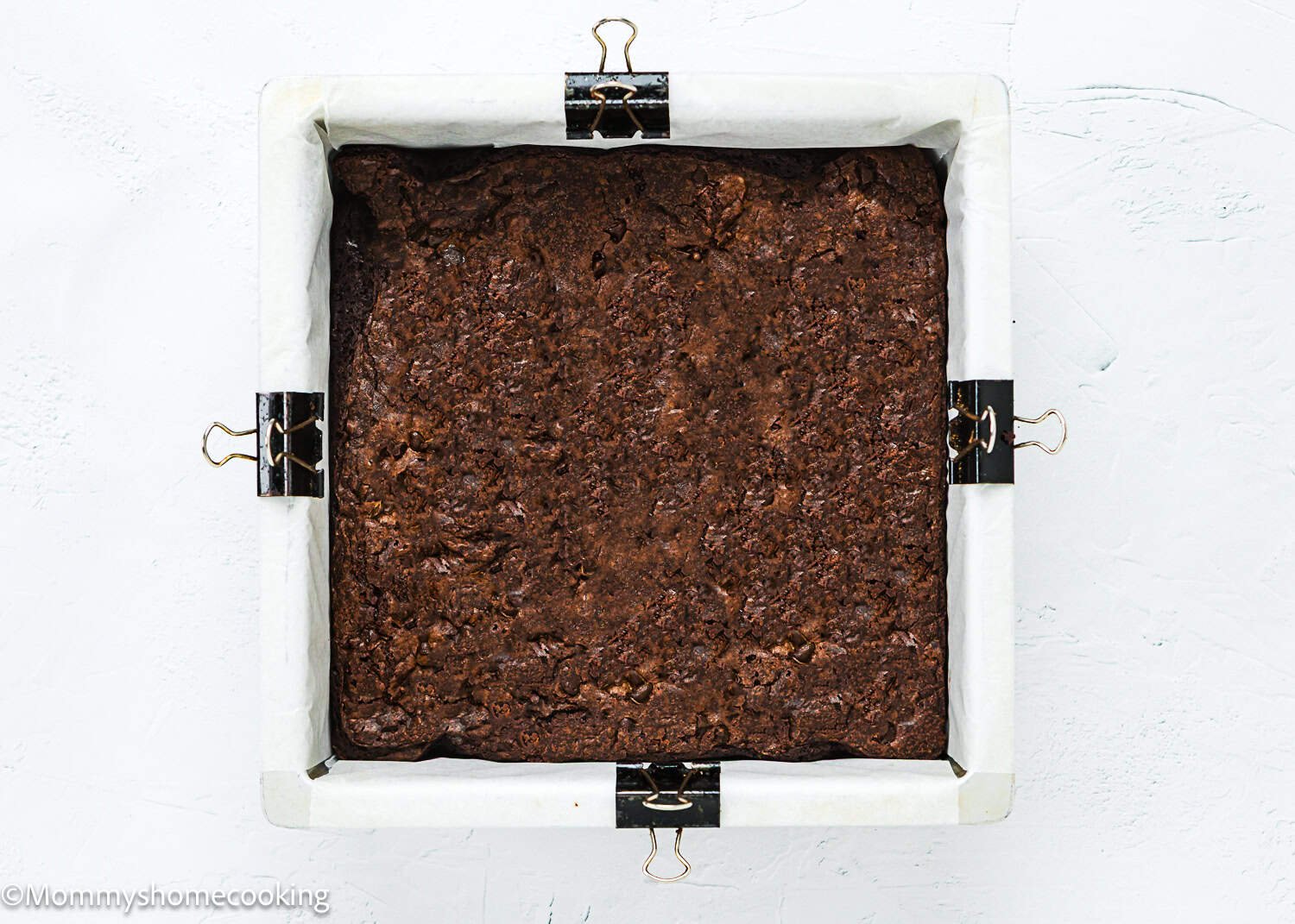 A square pan of baked brownies lined with parchment paper, secured at the edges with metal binder clips, shown from above on a white surface—perfect as a base for Eggless Ice Cream Cake.