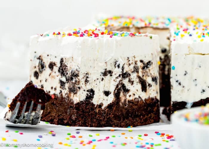 A close-up of a slice of eggless ice cream cake with a brownie base, topped with whipped cream and colorful sprinkles.