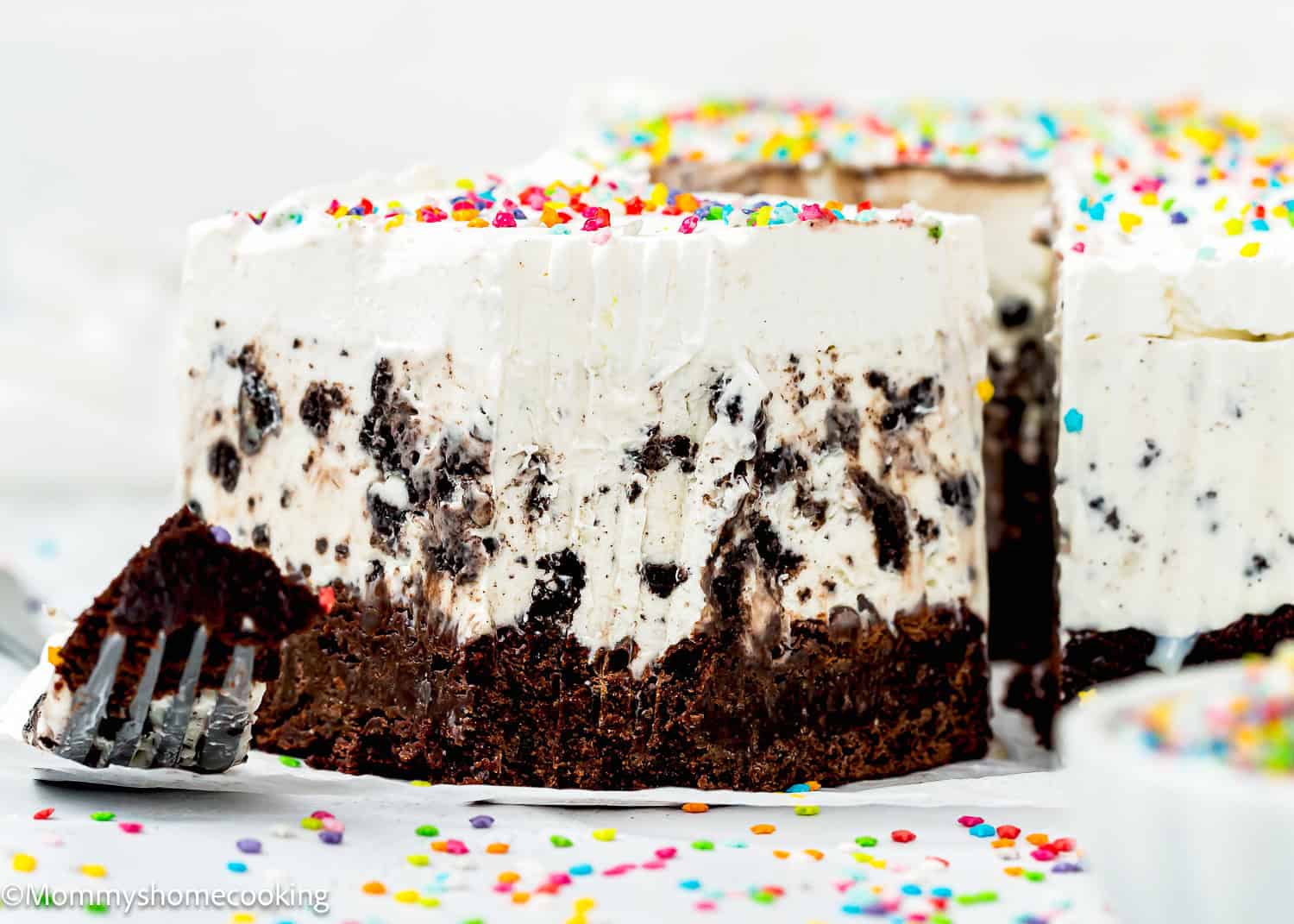 A close-up of a slice of eggless ice cream cake with a brownie base, topped with whipped cream and colorful sprinkles.