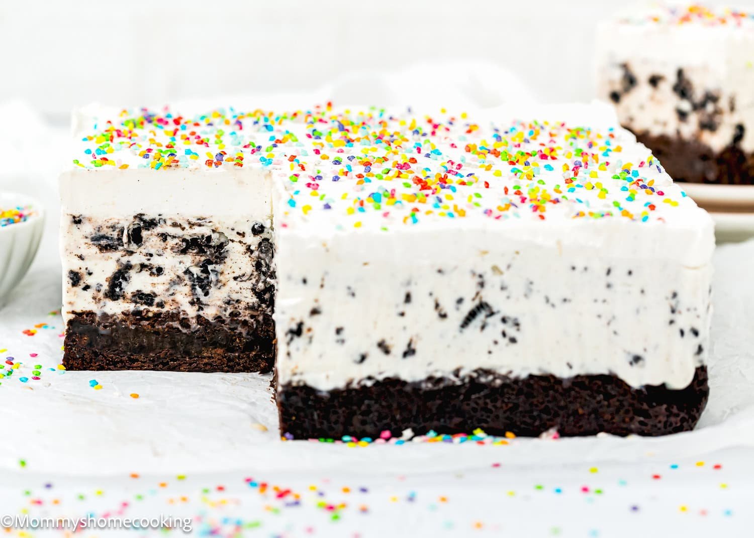 A rectangular Eggless Ice Cream Cake with a chocolate base, topped with whipped cream and colorful sprinkles, with one slice removed.