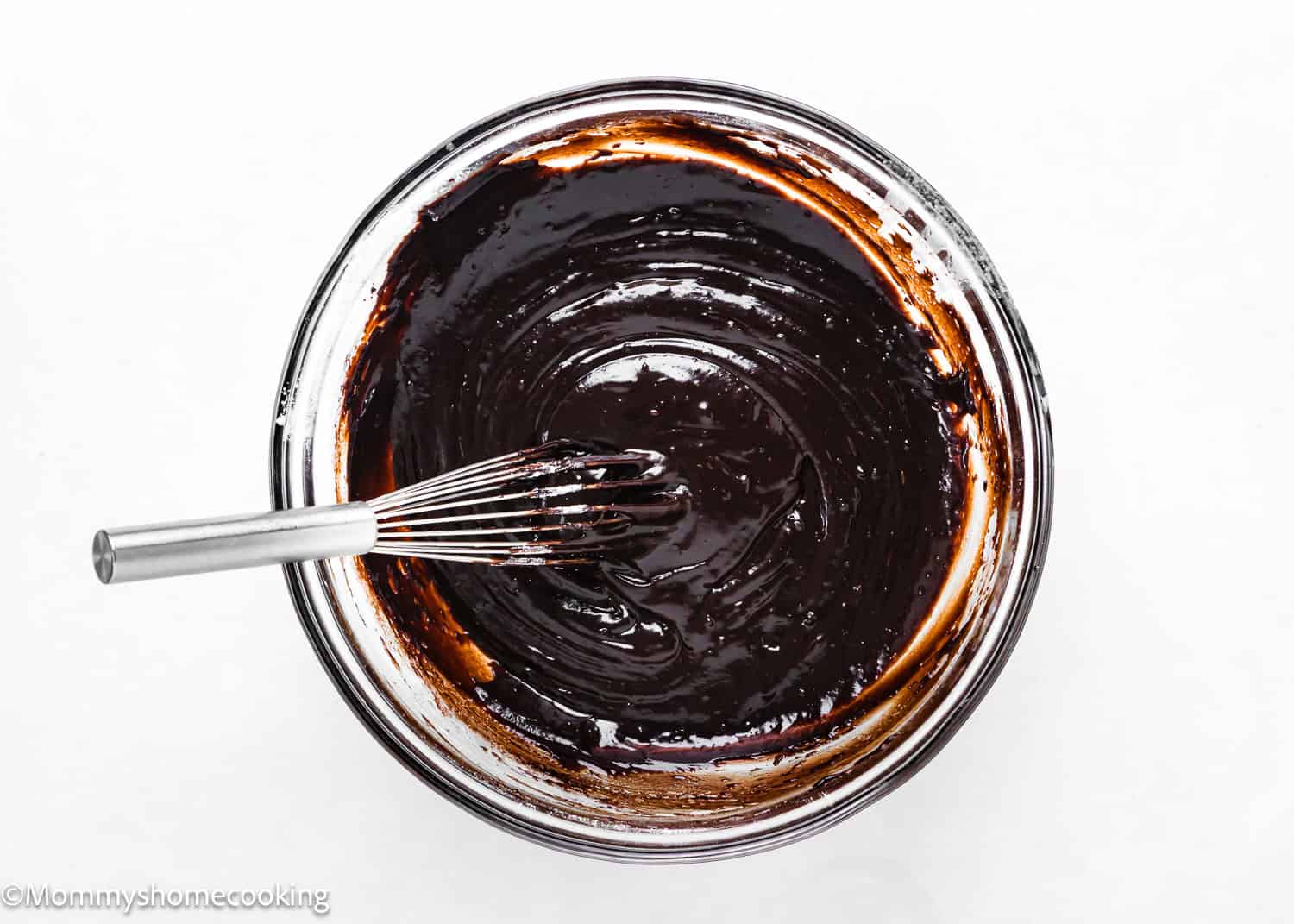 A metal whisk rests in a glass bowl filled with dark chocolate batter for Eggless Ice Cream Cake, viewed from above on a white surface.