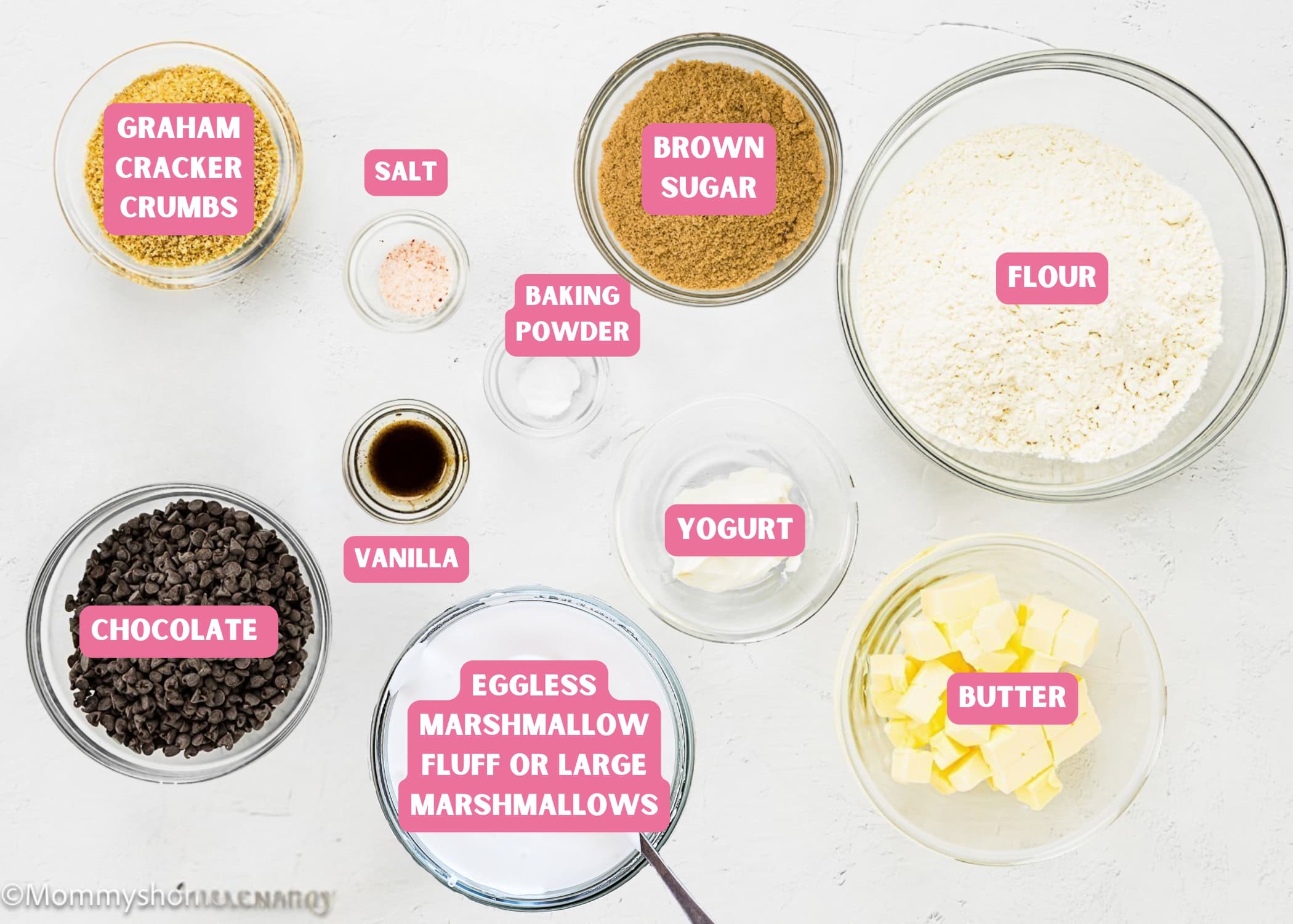 Various baking ingredients in clear bowls labeled as graham cracker crumbs, salt, brown sugar, flour, baking powder, vanilla, yogurt, butter, chocolate, and eggless marshmallow fluff for making Eggless S’mores Cookie Bars.