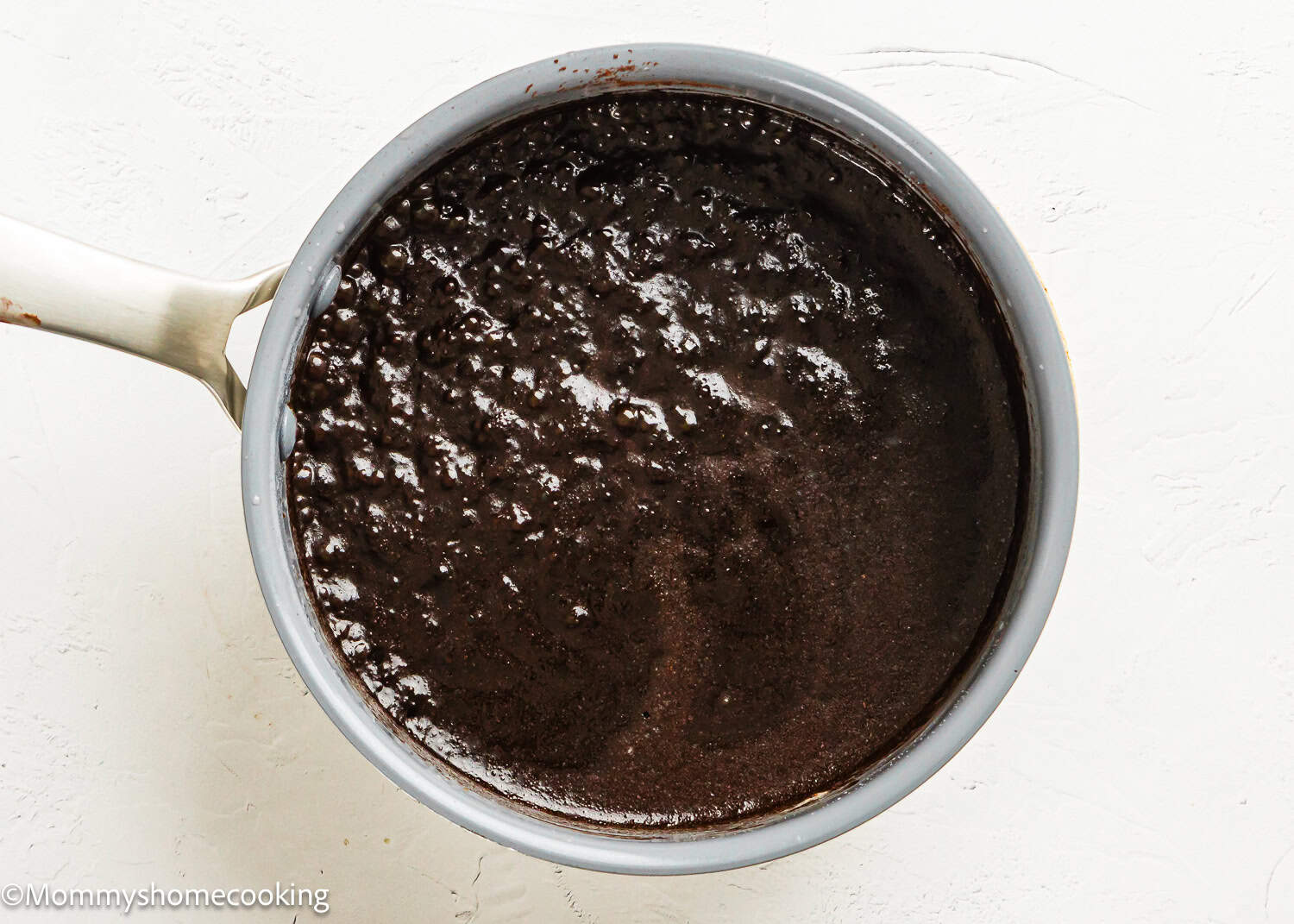 A saucepan filled with thick, bubbling Eggless Texas Sheet Cake chocolate mixture sits on a white surface.