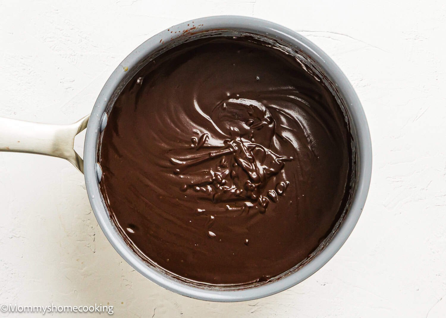 A saucepan filled with thick, glossy Eggless Texas Sheet Cake chocolate mixture on a white surface.