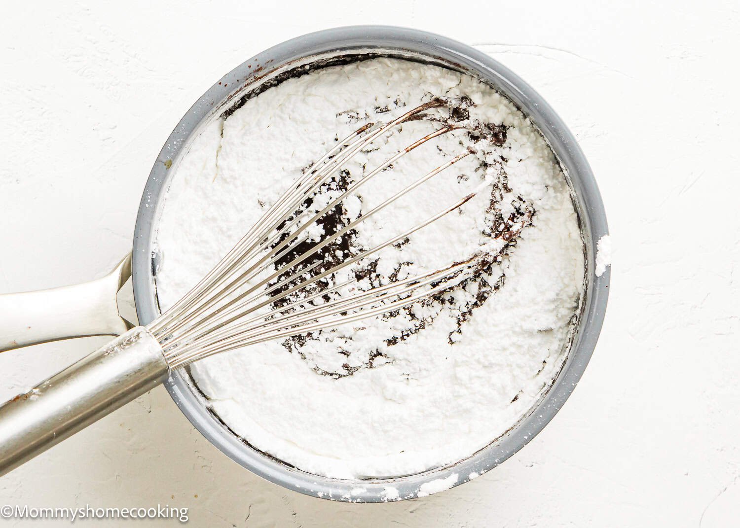 A metal whisk rests in a gray bowl filled with powdered sugar, partially mixed to reveal a dark liquid beneath—a key step in making Eggless Texas Sheet Cake.