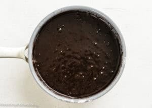 A saucepan filled with smooth, dark chocolate batter for Eggless Texas Sheet Cake sits on a white surface.