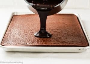 An Eggless Texas Sheet Cake is being topped with poured chocolate frosting.