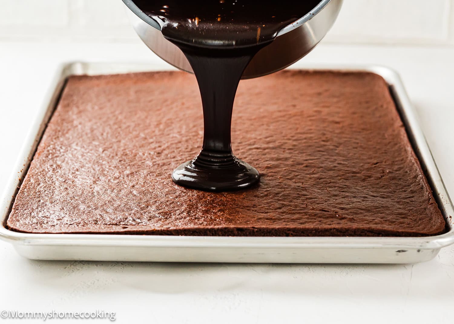 An Eggless Texas Sheet Cake is being topped with poured chocolate frosting.