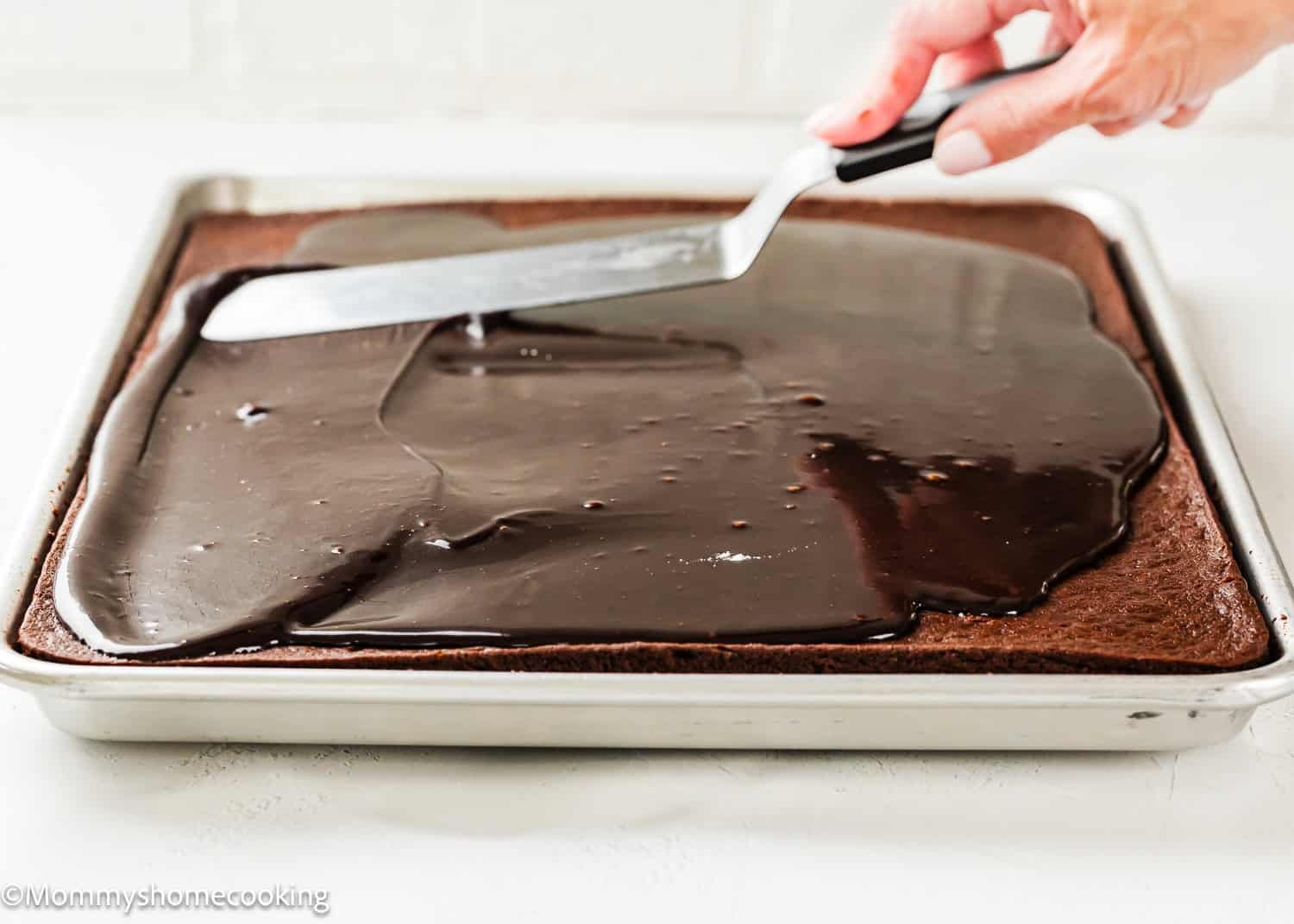 A hand uses an offset spatula to spread chocolate frosting over a large rectangular Eggless Texas Sheet Cake in a baking pan.
