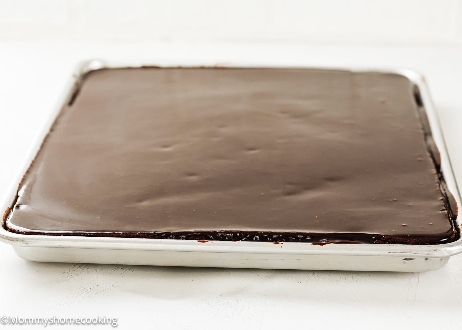 A large rectangular Eggless Texas Sheet Cake with a smooth layer of chocolate icing in a metal baking pan.