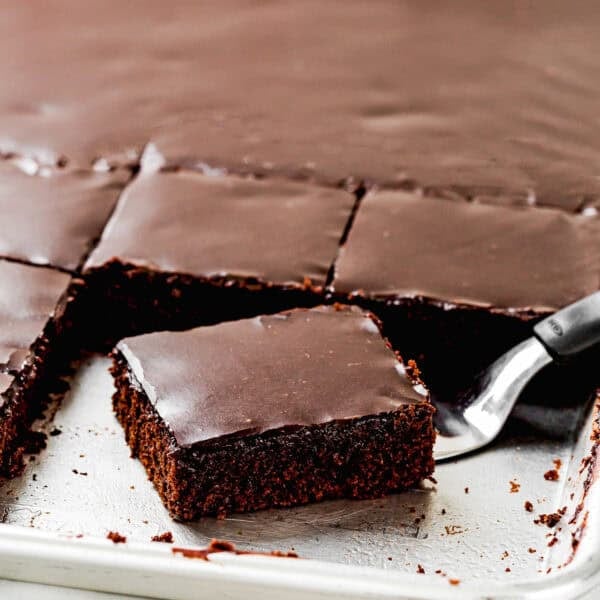 A metal spatula lifts a square piece of Eggless Texas Sheet Cake with glossy chocolate icing from a baking pan.