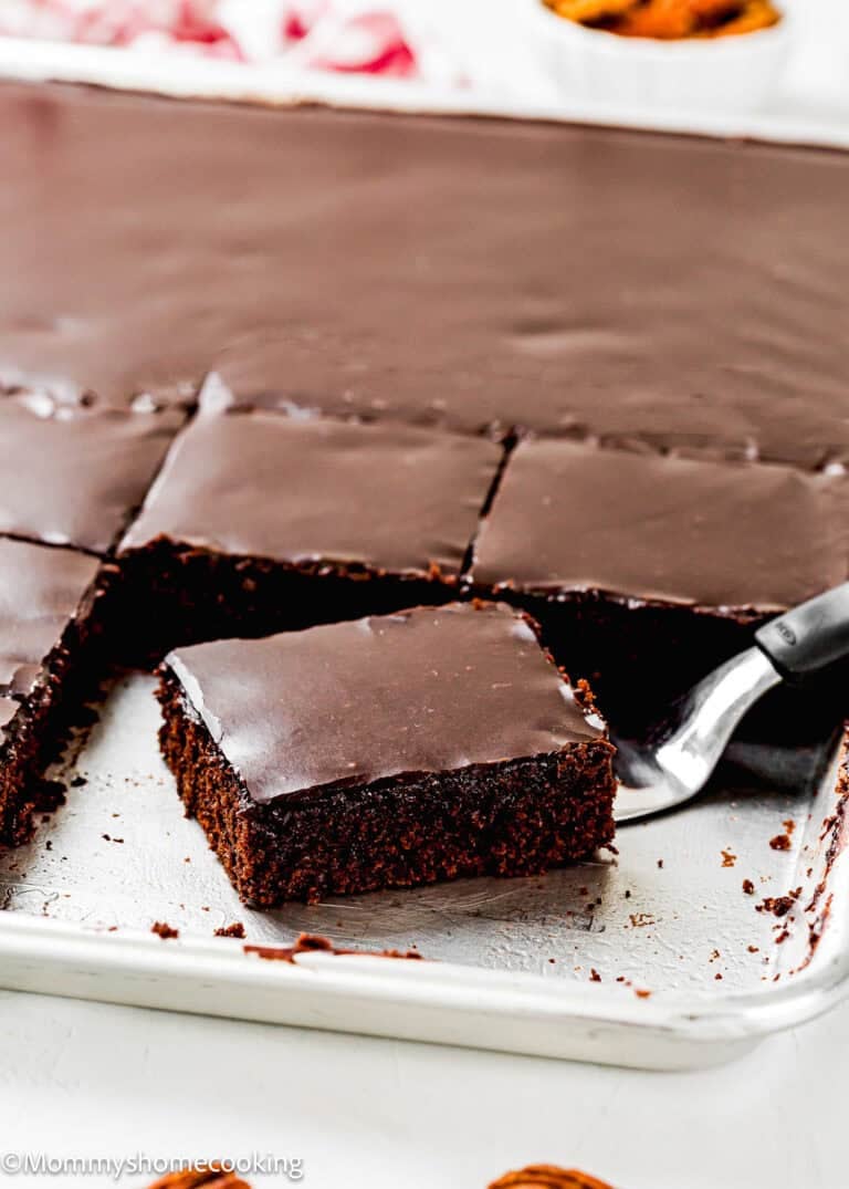 A metal spatula lifts a square piece of Eggless Texas Sheet Cake with glossy chocolate icing from a baking pan.
