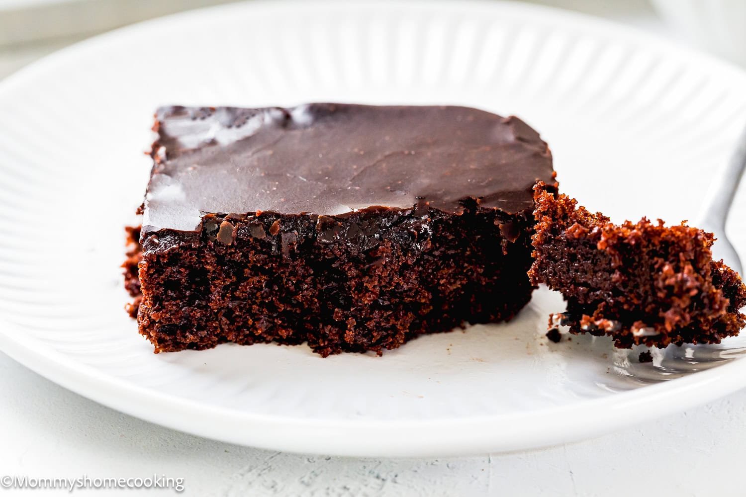 A slice of Eggless Texas Sheet Cake with glossy chocolate frosting on a white plate, with a piece cut off and placed beside it.