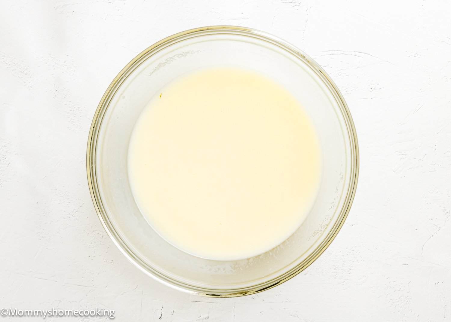 A glass bowl filled with a light yellow liquid mixture, perfect for making Eggless Texas Sheet Cake, sits on a white textured surface.