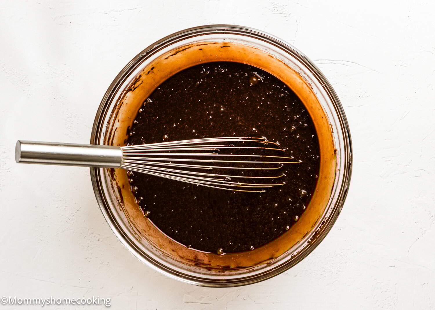 A glass bowl filled with Eggless Texas Sheet Cake chocolate batter and a metal whisk on a white surface.