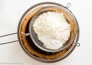 A metal sifter filled with flour, baking powder, and salt sits over a glass bowl containing chocolate batter for Eggless Texas Sheet Cake.