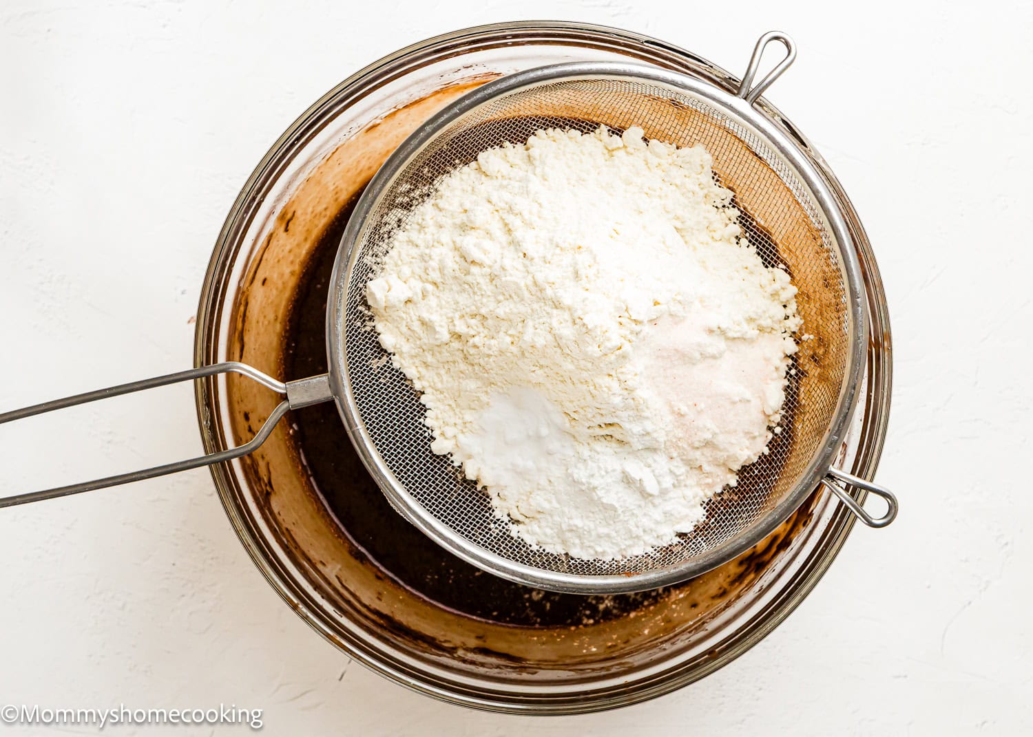A metal sifter filled with flour, baking powder, and salt sits over a glass bowl containing chocolate batter for Eggless Texas Sheet Cake.