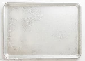 A silver baking sheet is lightly sprayed with oil, ready for preparing Eggless Texas Sheet Cake, and rests on a white surface.
