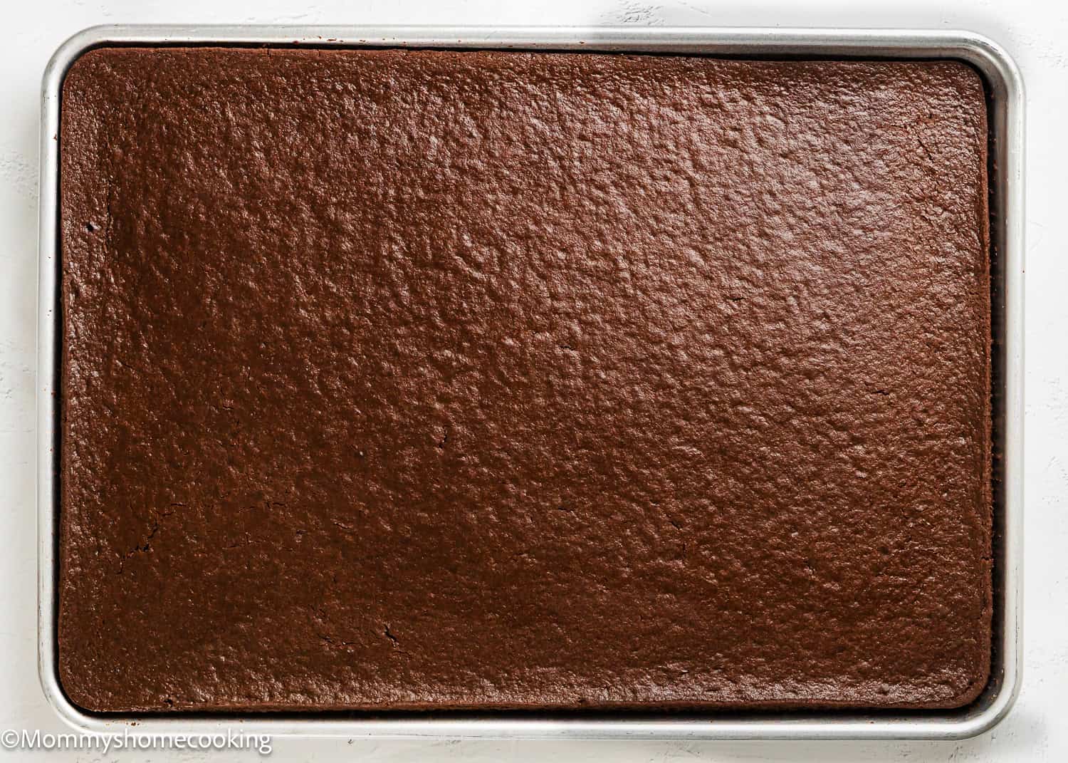 A sheet pan filled with a large, uncut, baked Eggless Texas Sheet Cake rests on a white surface.