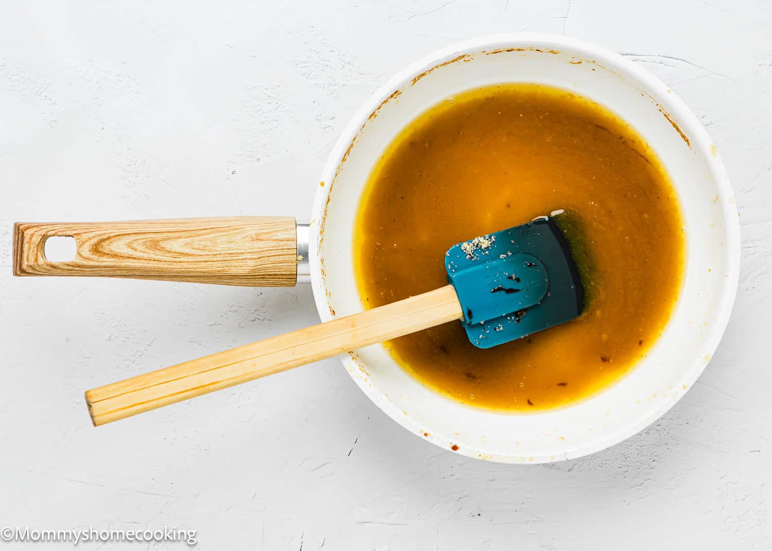 A white saucepan with a light wooden handle contains a butter mixture, perfect for an Eggless Upside-Down Peach Cake, with a blue spatula resting inside.