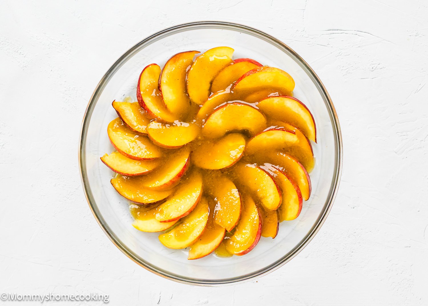 A glass bowl containing neatly arranged peach slices topped with a sugar glaze sits on a white textured surface, perfect for serving alongside Eggless Upside-Down Peach Cake.