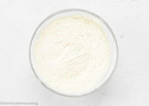 A glass bowl filled with a fine white powder, possibly flour or a baking mix for Eggless Upside-Down Peach Cake, placed on a white surface.