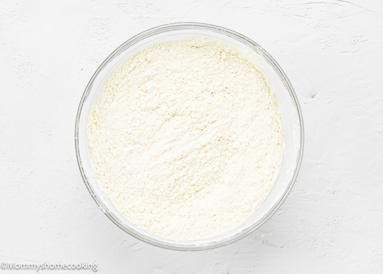 A glass bowl filled with dry ingredients for Eggless Upside-Down Peach Cake, placed on a white surface.