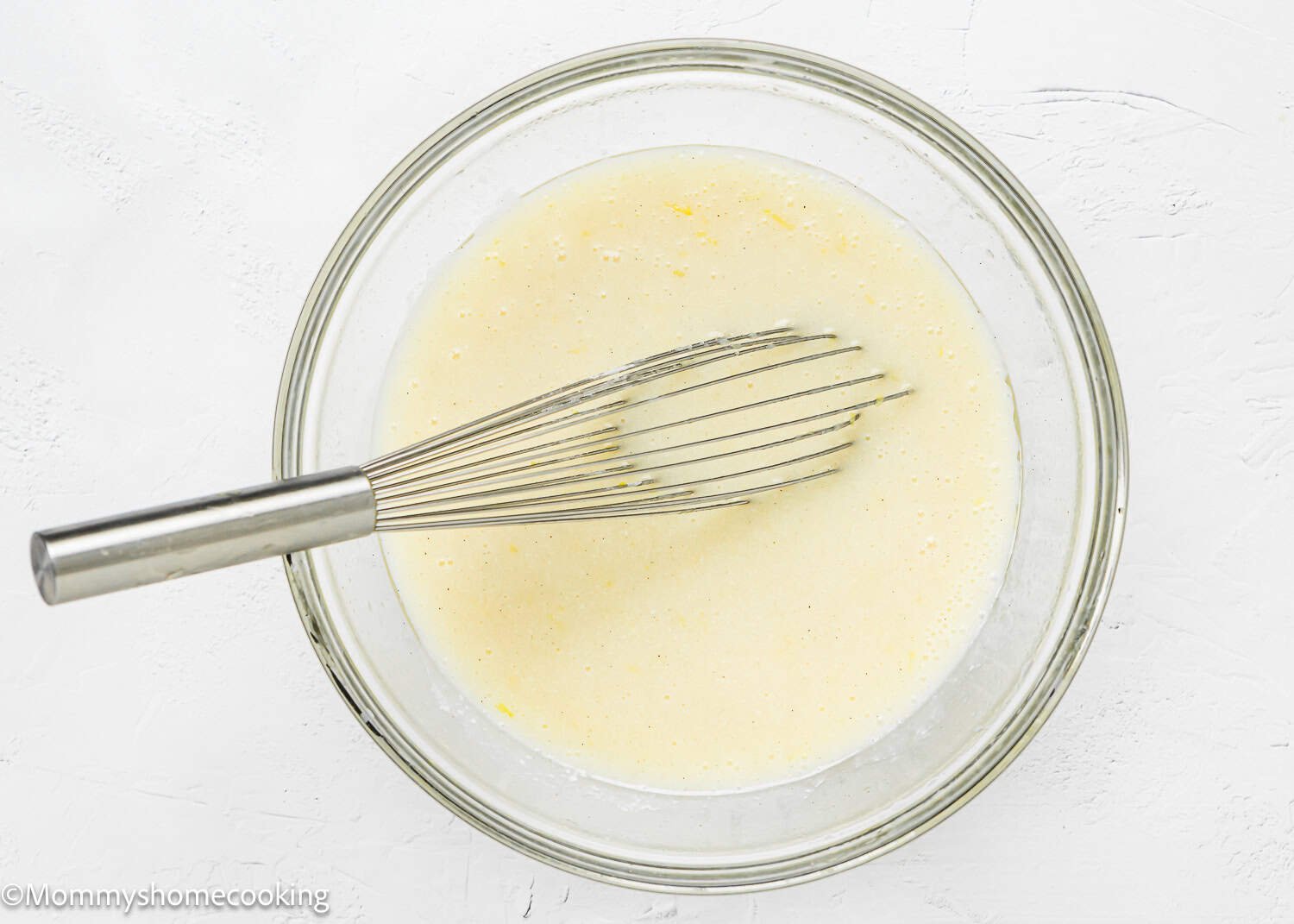 A metal whisk rests in a glass bowl filled with wet ingredients for an Eggless Upside-Down Peach Cake on a white surface.