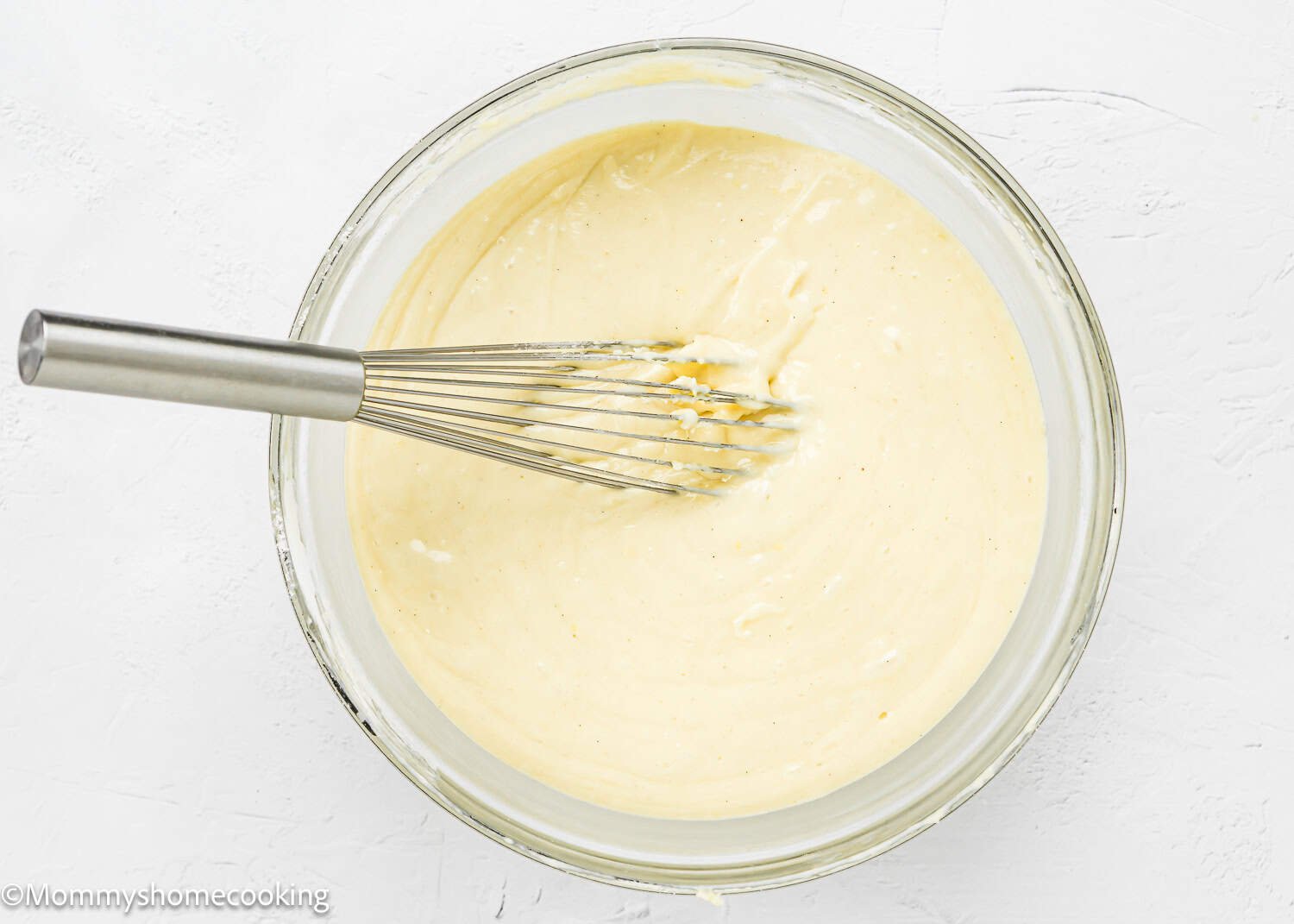 A metal whisk rests in a glass bowl filled with smooth, pale yellow Eggless Upside-Down Peach Cake batter on a white surface.
