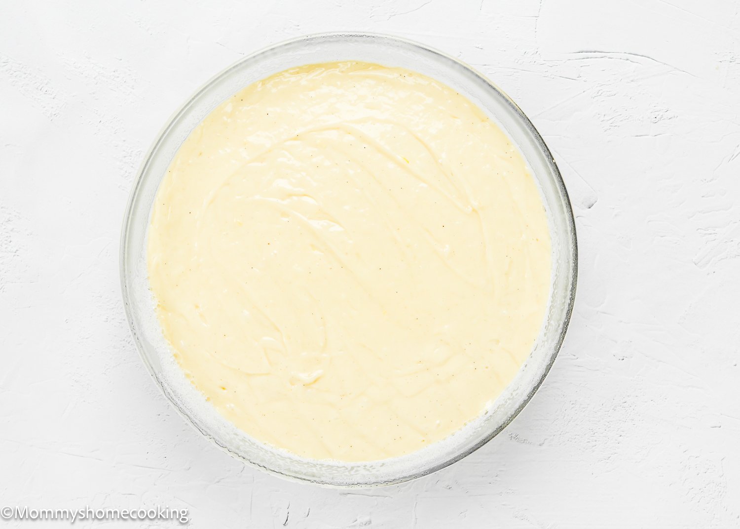 A glass bowl filled with smooth, creamy batter for Eggless Upside-Down Peach Cake sits on a white textured surface.