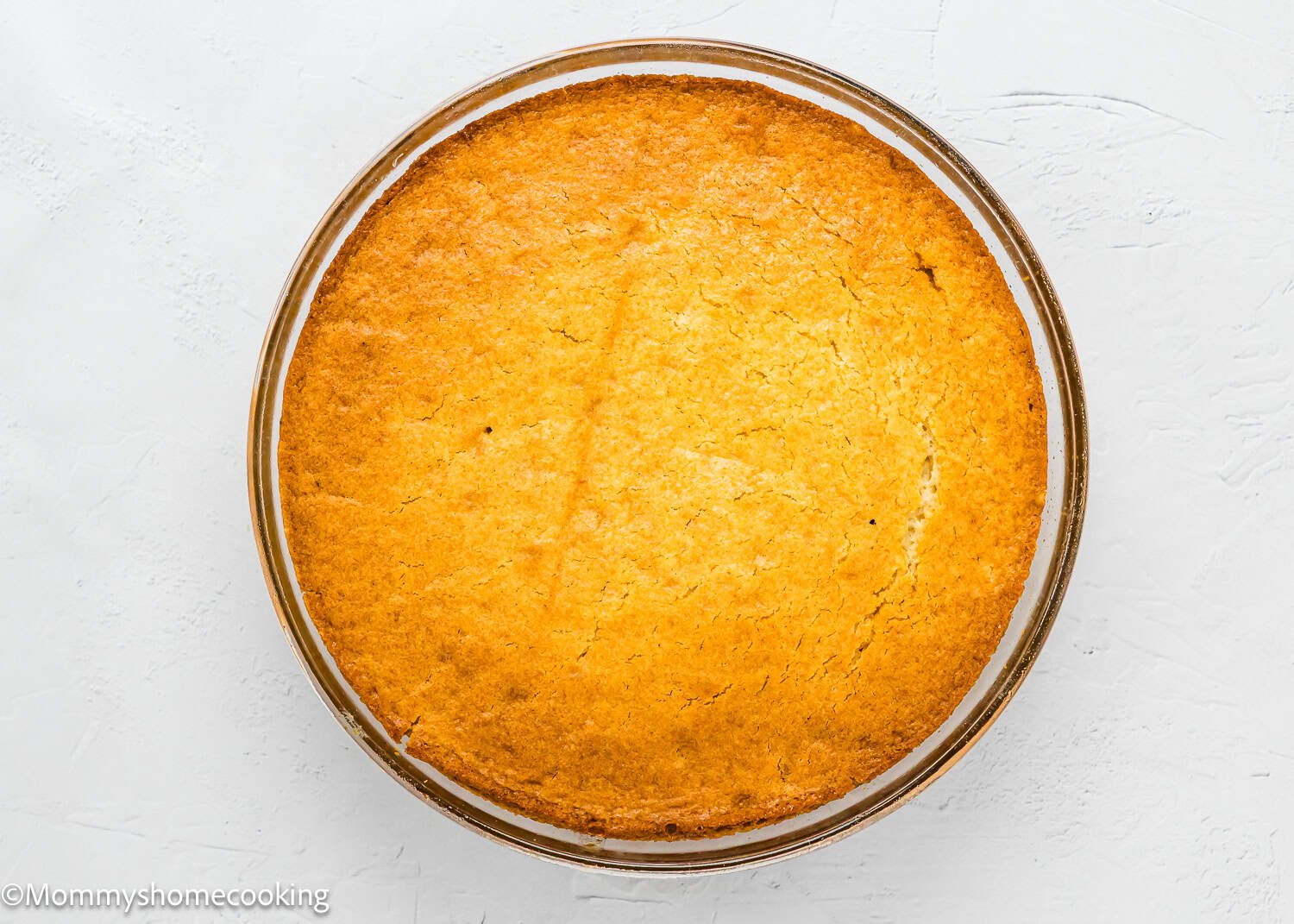 An Eggless Upside-Down Peach Cake, round and golden-brown, sits in a glass baking dish on a white textured surface.