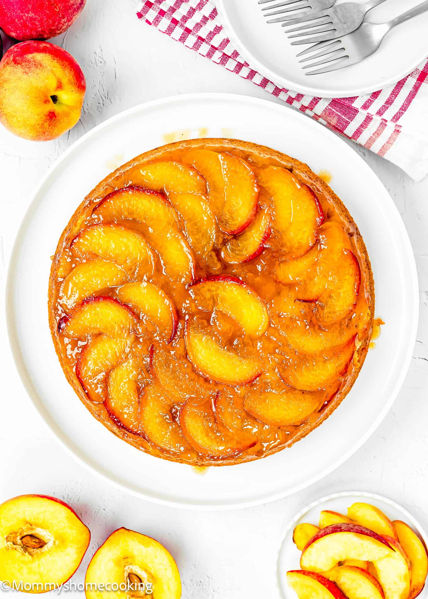 An Eggless Upside-Down Peach Cake sits on a white plate, topped with glazed peach slices. Sliced peaches and whole peaches are arranged nearby on the white table.