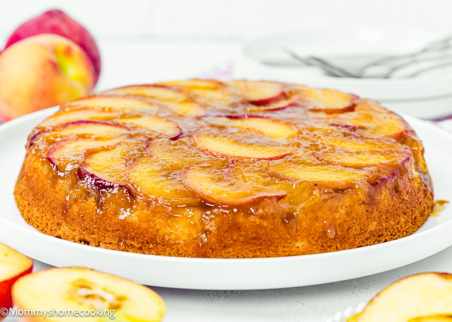 An Eggless Upside-Down Peach Cake with glazed peach slices sits on a white plate, surrounded by fresh peaches in the background.