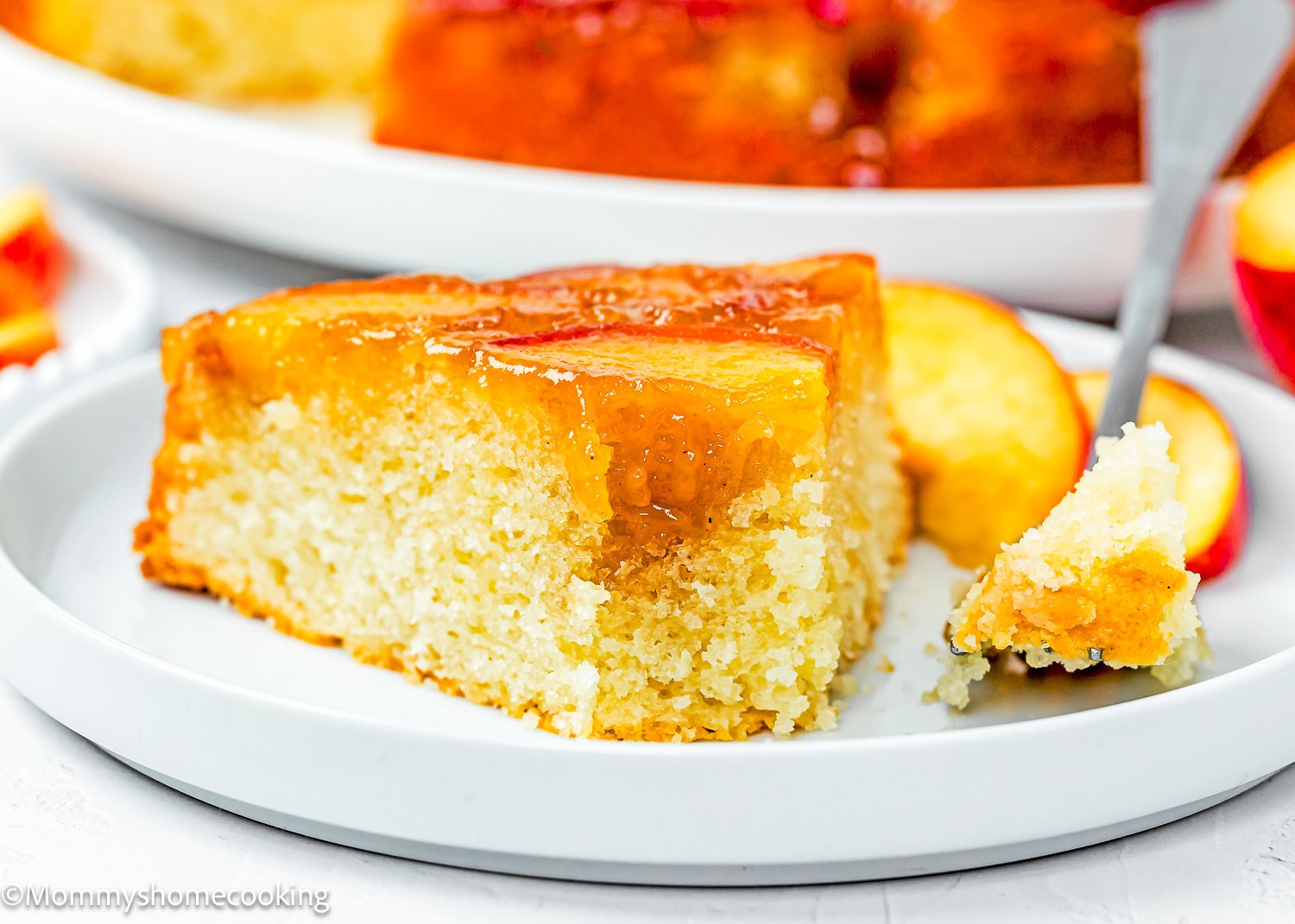 A slice of Eggless Upside-Down Peach Cake sits on a white plate with a fork holding a bite-sized piece.