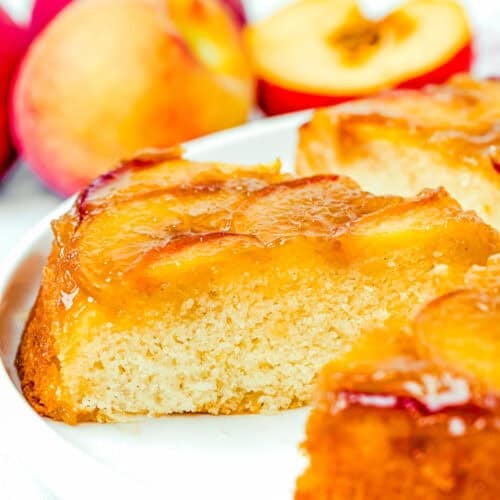 A close-up of an Eggless Upside-Down Peach Cake slice on a white plate, with fresh peaches in the background.