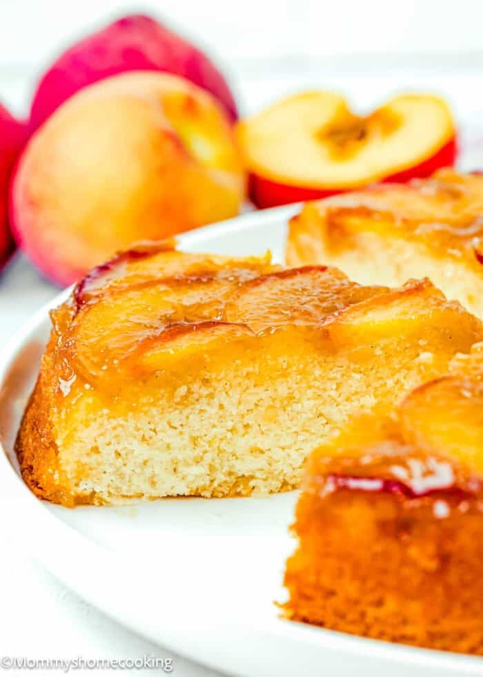 A close-up of an Eggless Upside-Down Peach Cake slice on a white plate, with fresh peaches in the background.