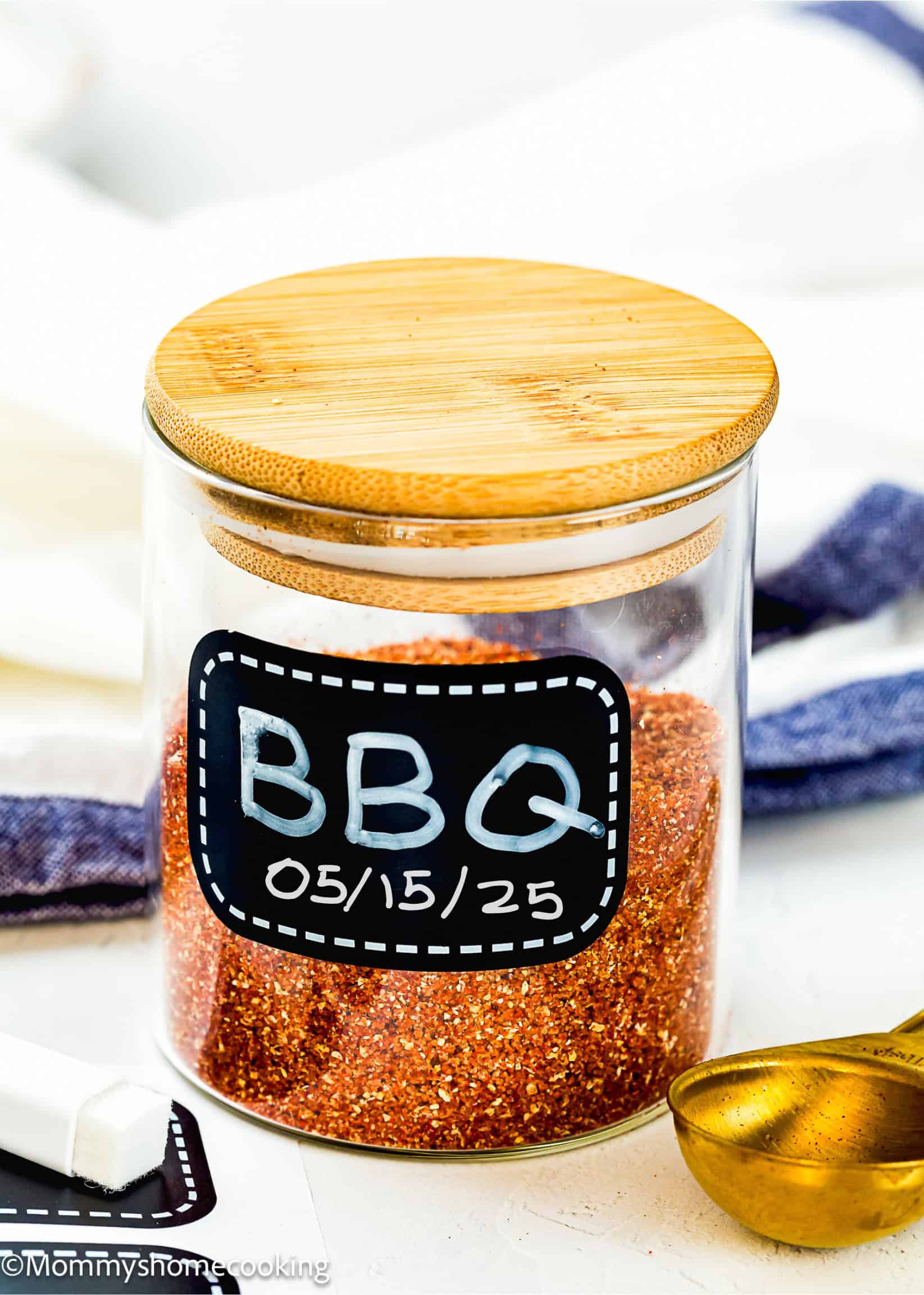 A glass jar with a wooden lid labeled "BBQ" contains a Homemade Barbecue Meat Rub. A gold measuring spoon and a piece of chalk lie nearby.