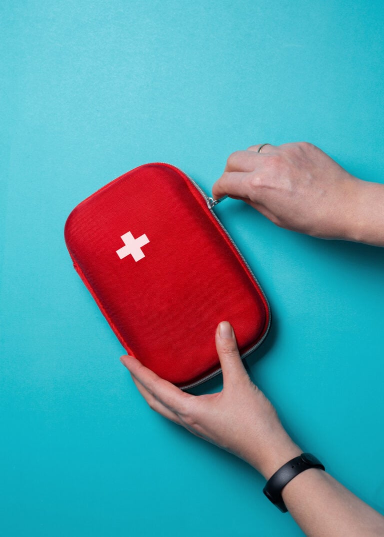 A person opens a red first aid kit with a white cross on the cover, set against a blue background—an essential step in learning how to build an emergency kit for food allergies.