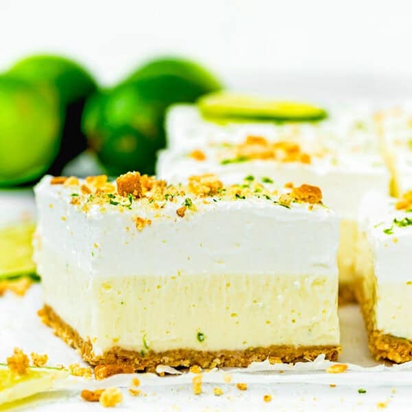 A close-up of No Bake Key Lime Pie Bars with a crumbly crust, creamy filling, and whipped topping, garnished with lime zest and cookie crumbs. Whole limes are blurred in the background.