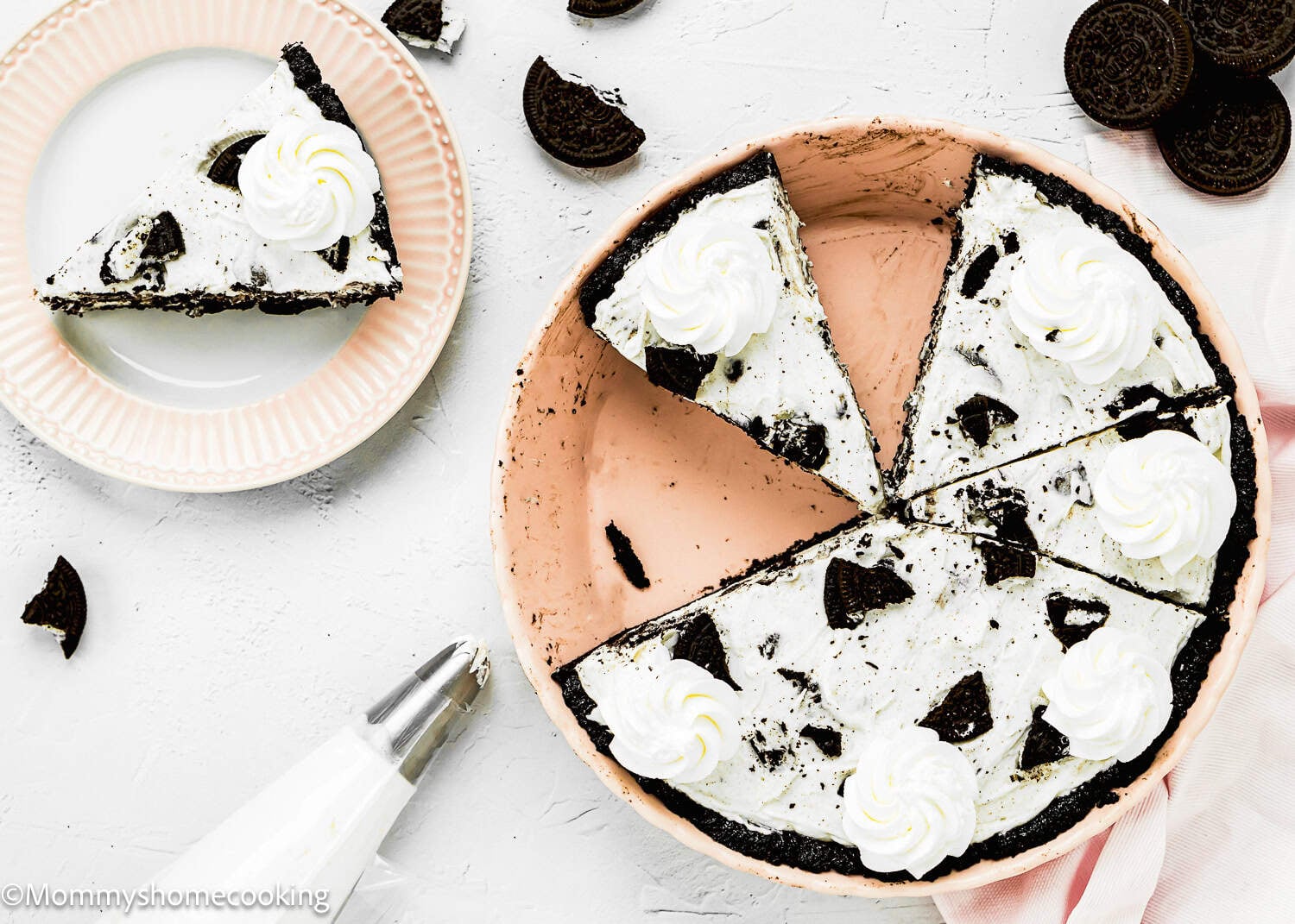A No Bake Oreo Cheesecake Pie with whipped cream topping is sliced, with one piece served on a plate. A piping bag and cookie pieces are nearby.