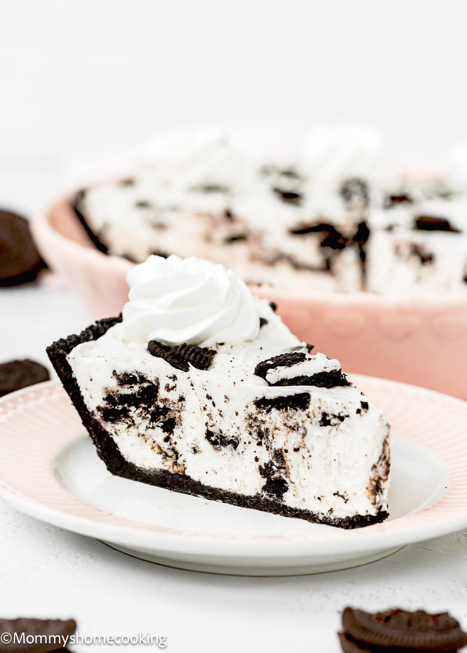 A slice of No Bake Oreo Cheesecake Pie with a chocolate crust and whipped cream topping sits on a plate, with the rest of the pie and cookie pieces in the background.