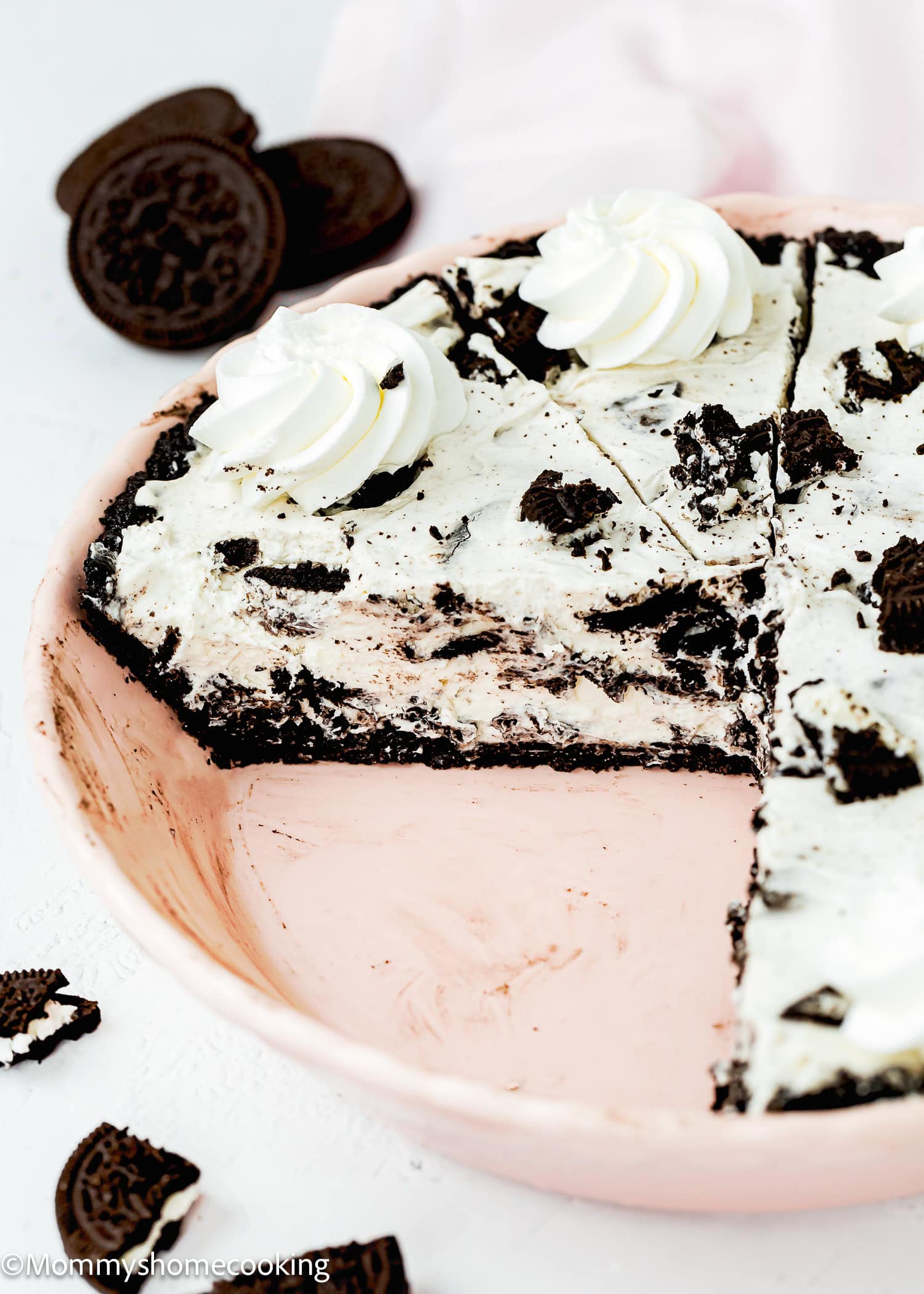 A partially sliced No Bake Oreo Cheesecake Pie with whipped cream topping in a pink pie dish, with Oreo cookies scattered nearby.