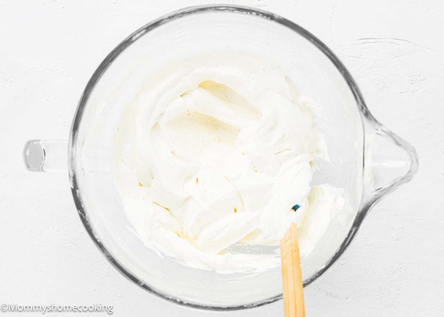 A glass mixing bowl containing a creamy white No Bake Oreo Cheesecake Pie mixture being stirred with a spatula on a white surface.