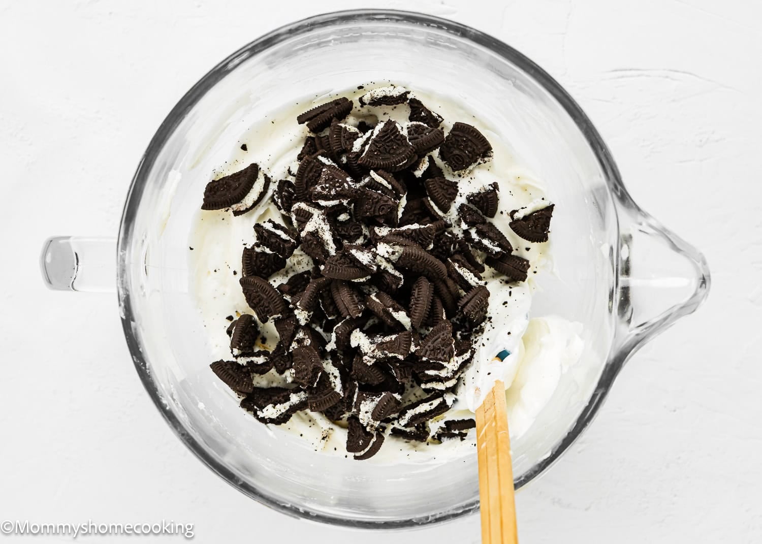 A glass bowl filled with creamy white No Bake Oreo Cheesecake Pie mixture and chopped chocolate sandwich cookies, with a wooden spatula resting inside.