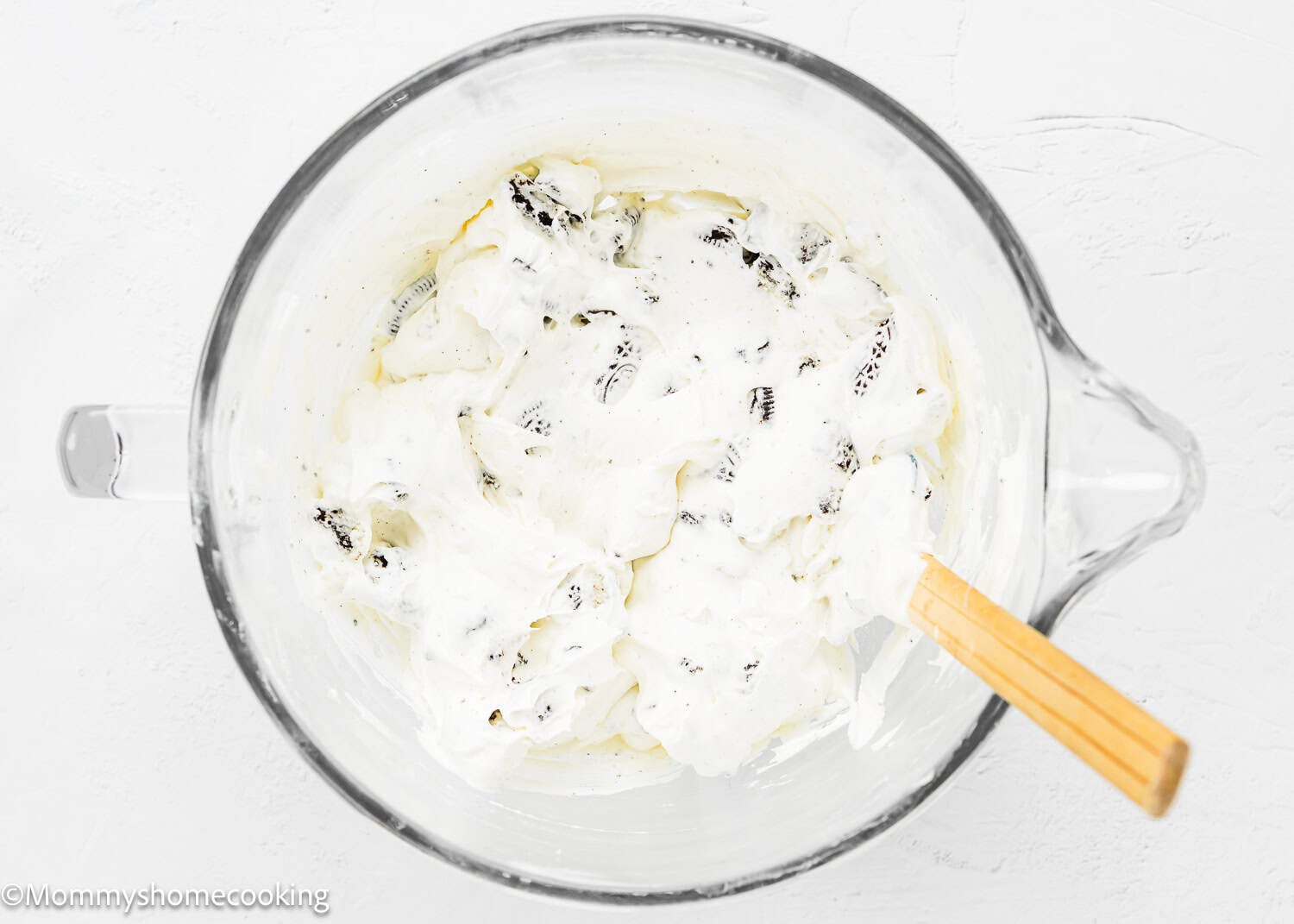 A glass mixing bowl filled with whipped cream and crushed cookies, being stirred with a wooden spatula on a white surface—perfect for making No Bake Oreo Cheesecake Pie.